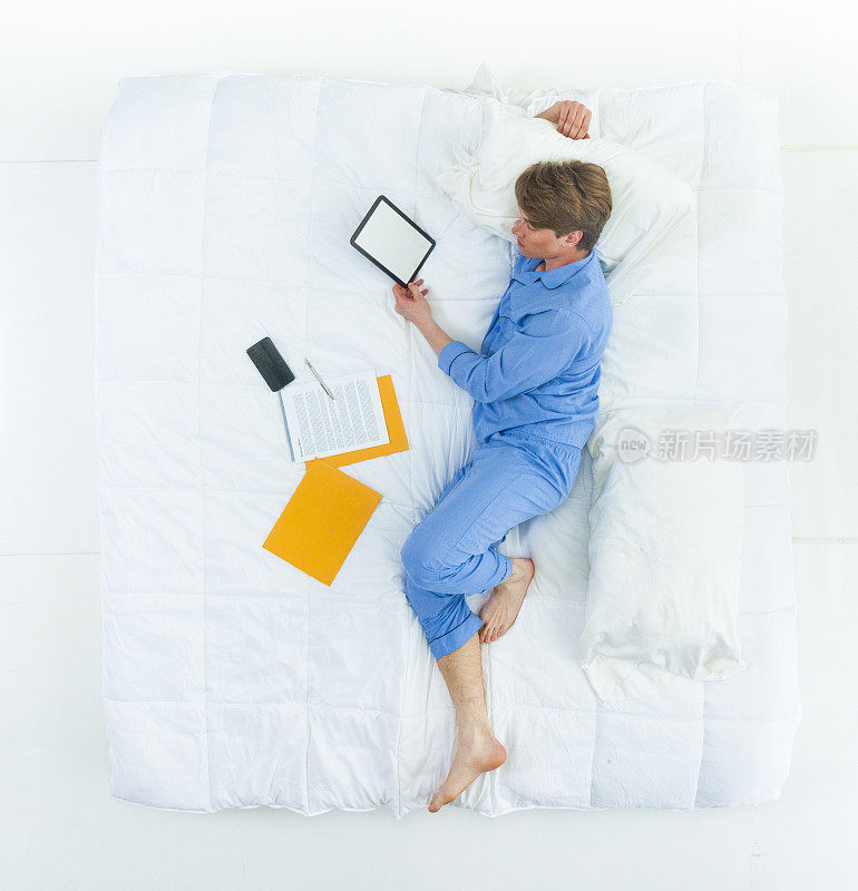 白人年轻男子/男性穿着睡衣在卧室的白色背景前睡觉/放松/休息，使用智能手机/数码平板电脑